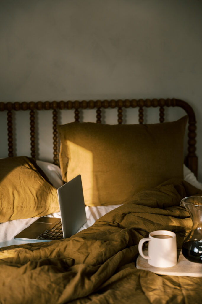 Soft morning light in a calm bedroom with neutral bedding, a laptop, and coffee, illustrating a home environment designed for gentle daily rhythms and smart home wellness. 