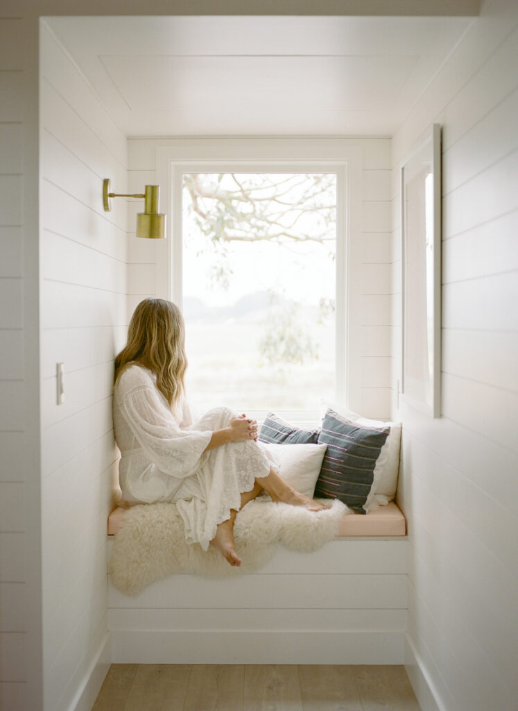 Woman in a white dress sitting on a cushioned window bench in a narrow white alcove, looking out a large window in soft natural light.