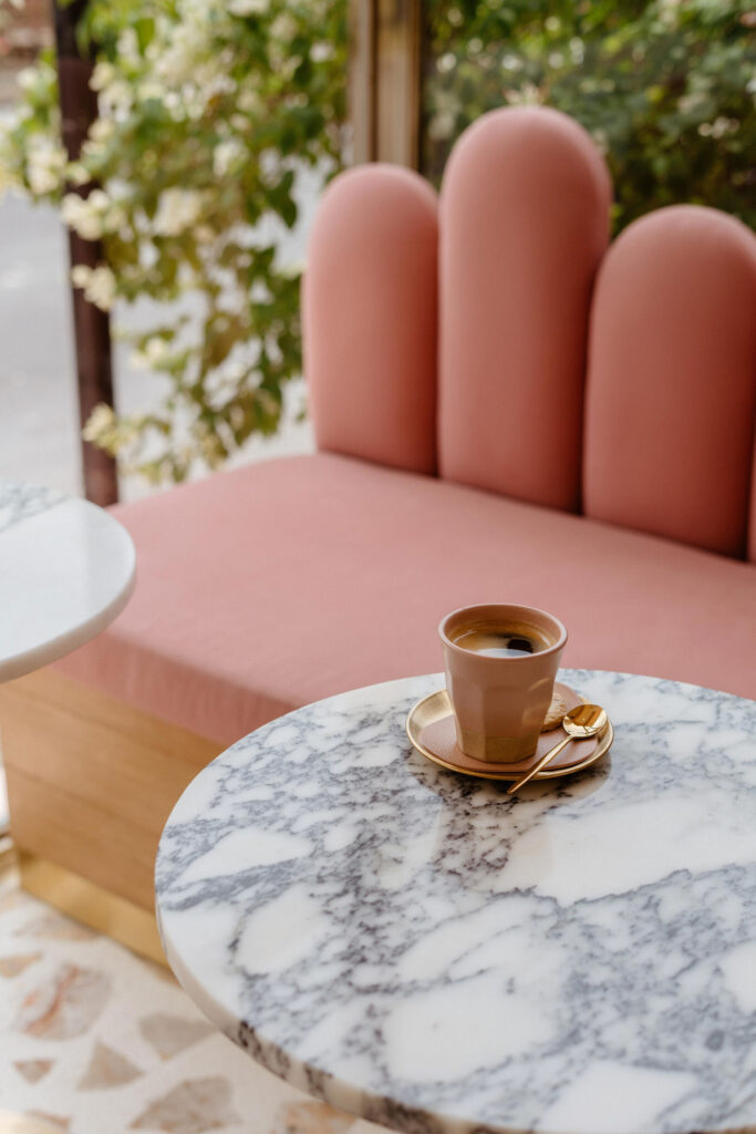 Cup of espresso on a marble café table in front of a blush pink upholstered banquette and leafy window view.
