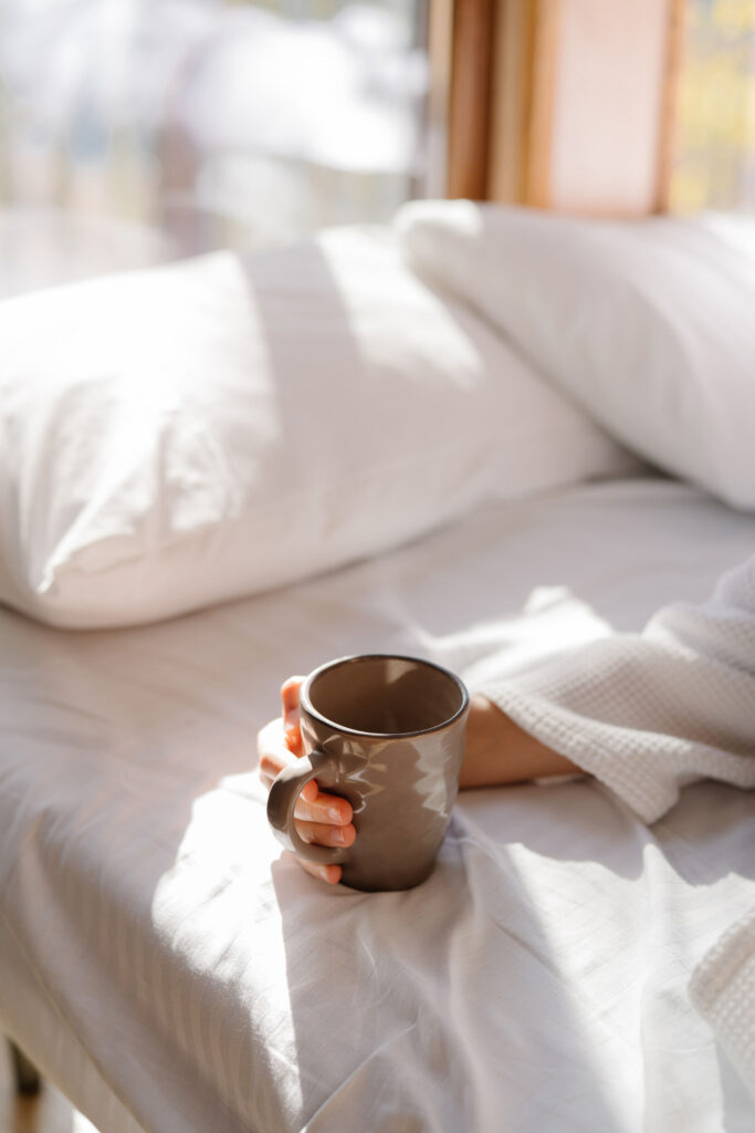 Morning sunlight across white bedding as someone relaxes in bed holding a coffee mug.