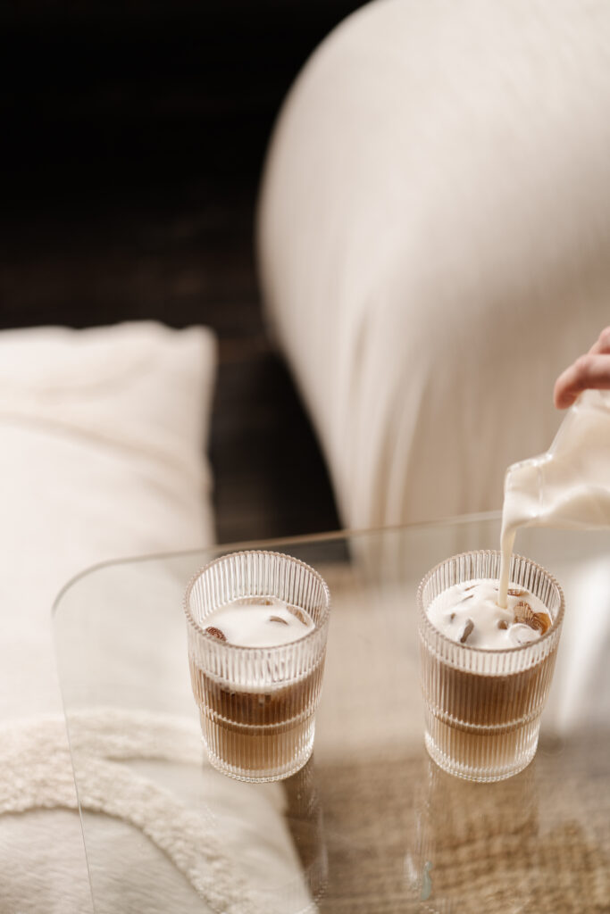 Milk being poured into two iced coffees on a glass bedside table beside a softly lit bed.