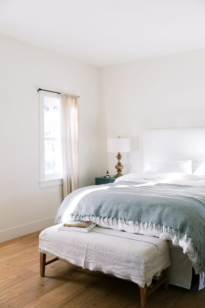 Bright, serene bedroom with white bedding, a soft blue throw, warm wood floors, and natural light streaming through a window.
