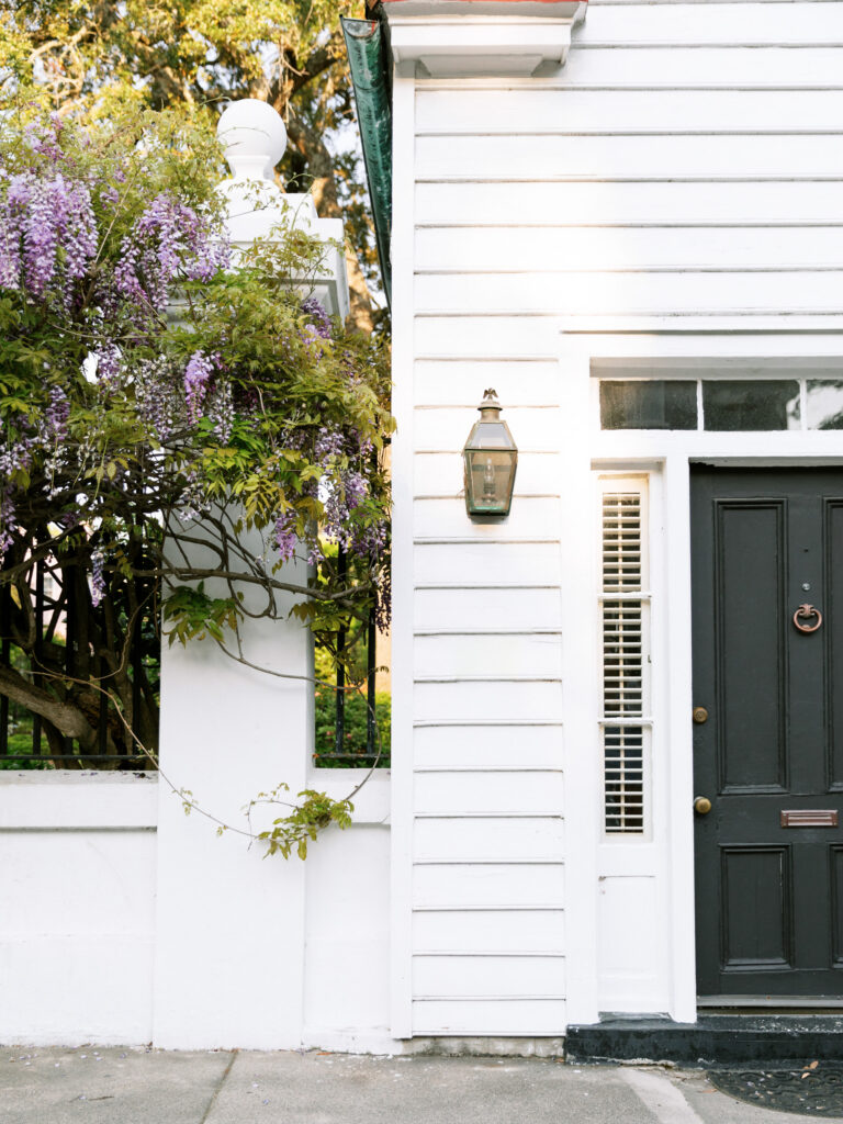 White house exterior with black front door, classic lantern, and blooming purple wisteria climbing beside the entry.